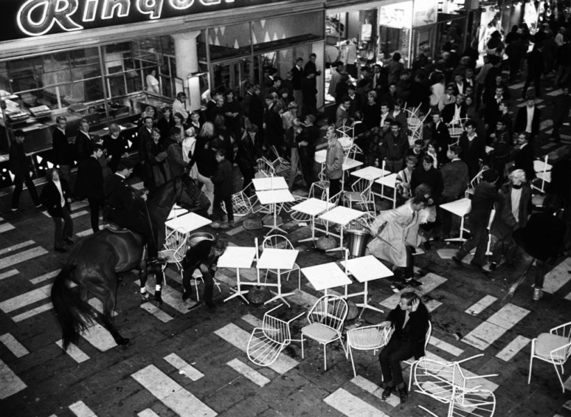 Uniformerad polis på häst tar sig in i folkmassorna bland människor, kafébord och stolar under de så kallade Hötorgskravallerna 1965.