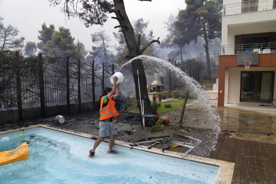 En man slänger vatten från en simbassäng medan elden närmar sig hans hus norr om Aten.