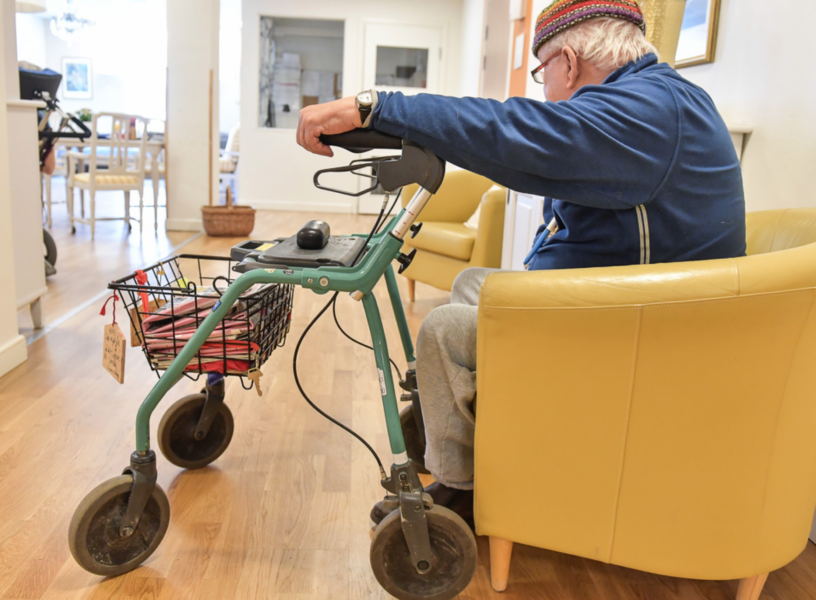 En äldre man med rollator sitter i en korridor på ett vård- och omsorgsboende.
