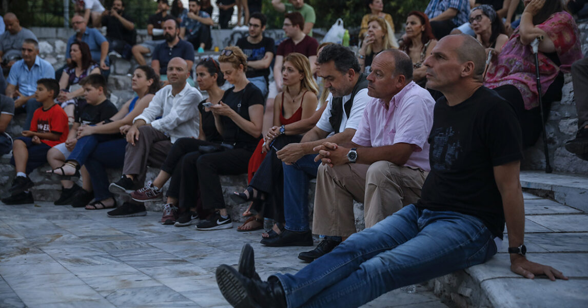 Människor på en trappa lyssnar på någon. Närmast kameran sitter Yanis Varoufakis.