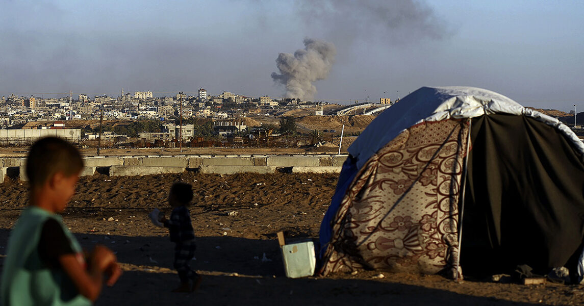 En pojke står vid ett tält och ser på avstånd hus i staden Rafah bombas.
