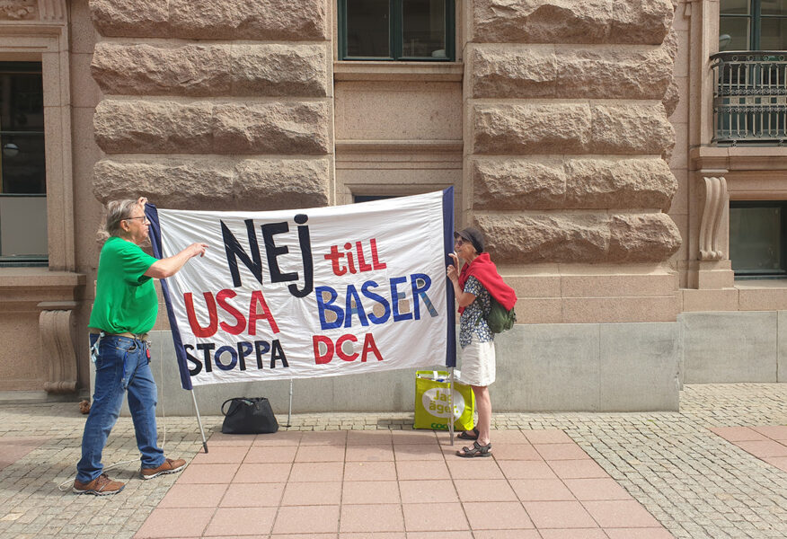 Två personer håller en banderoll med texten Nej till USAbaser Stoppa DCA