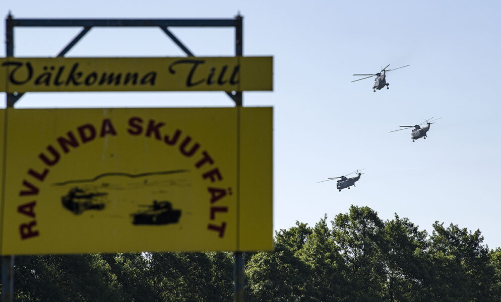 Skylt med texten Välkomna till Ravlunda skjutfält, tre helikoptrar flyger i bakgrunden
