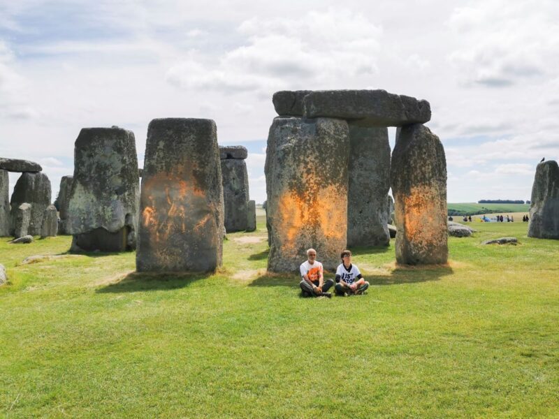 två personer sitter på gräset framför ett vandaliserat stonehenge