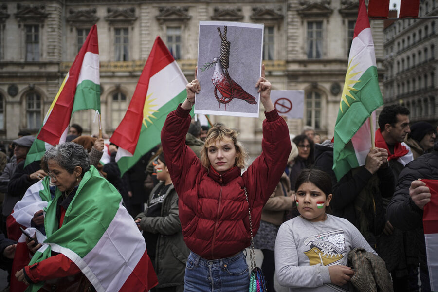 Personer vid demonstration, iranska flaggor, en kvinna håller upp en bild med en blodig duva i en hängsnara.