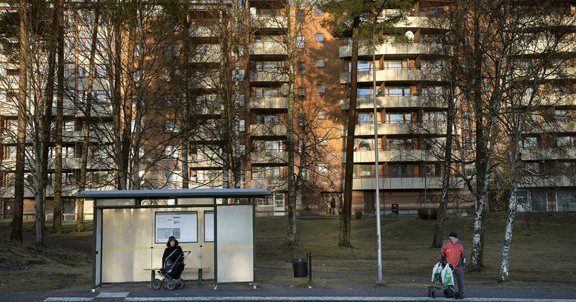 En busshållplats framför ett stort hyreshus och lite tallar.