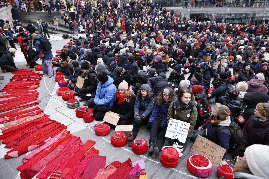 Rebellmammor i sittande demonstration med röda halsdukar utrullade på marken.