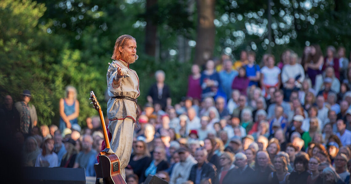 En skådespelare med gitarr och publik i en park på sommaren.