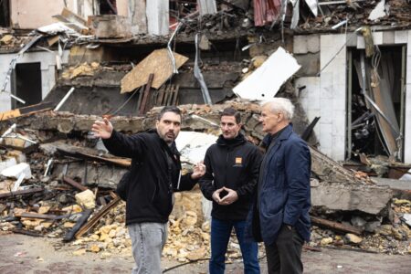 Jan Egeland och två medarbetare på NRC står framför ett sönderbombat hus.