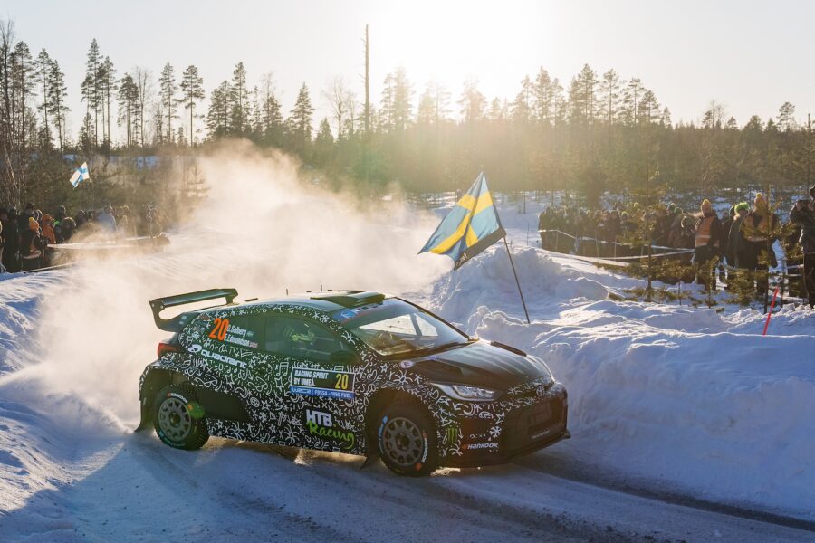 Rallybil kör i snön med åskådare längs vägen.