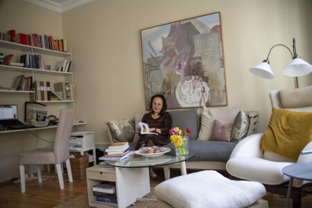 Anita Goldman sittandes i soffan i sin lägenhet, bokhylla, skrivbord, stor tavla på väggen bakom henne