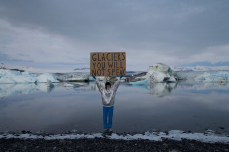 Nastja Säde Rönkköhåller står i islandskap, håller upp en skylt med texten ”Glaciers, you will not speak”.