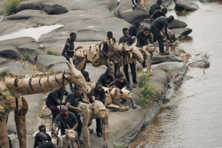 Teaterdockor föreställande djur i naturlig storlek står vid en flodbank tillsammans med ett antal personer.