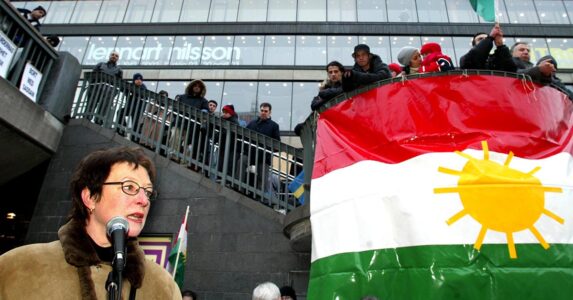 En kvinna talar framför en kurdisk flagga.