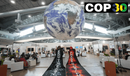 Aktivister med långa mantlar står med ryggen mot betraktaren, budskap om att stoppa användningen av fossila bränslen. Stor jordglob hänger i taket.