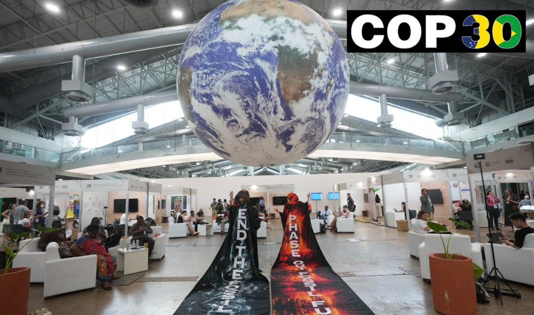 Aktivister med långa mantlar står med ryggen mot betraktaren, budskap om att stoppa användningen av fossila bränslen. Stor jordglob hänger i taket.