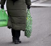 En kvinna med grön tygkasse och grön plastpåse i var hand.